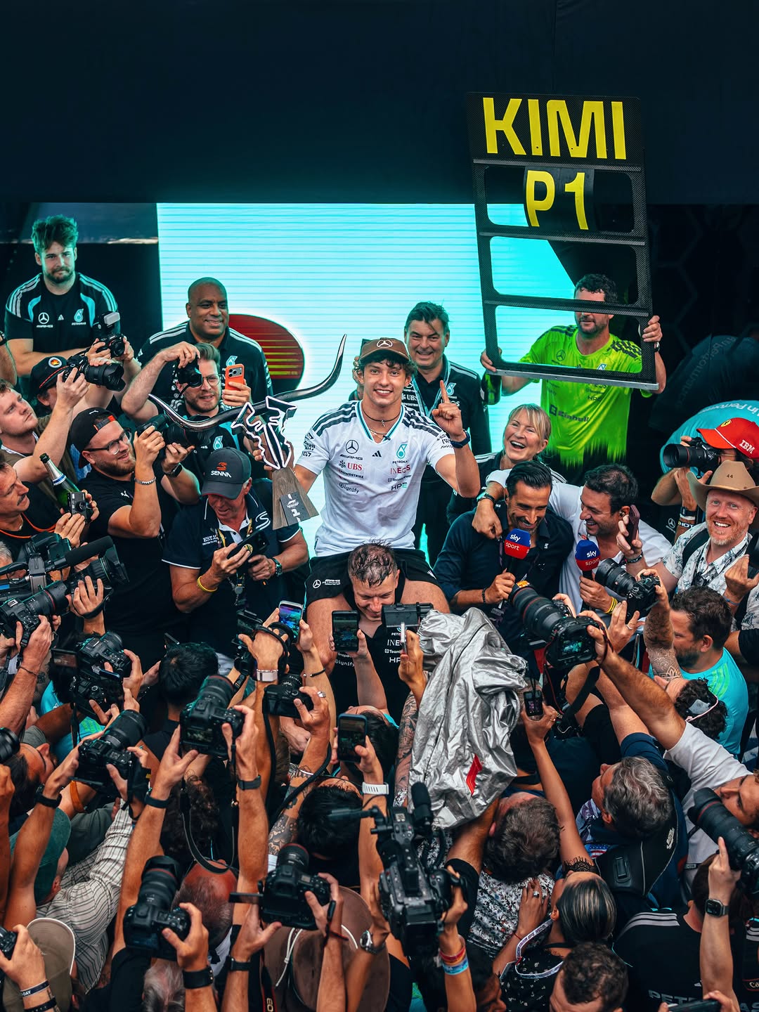 Kimi Antonelli, holding his Austin winner's trophy, is lifted by the Mercedes team under a KIMI P1 pit board after his ninth win of the season.