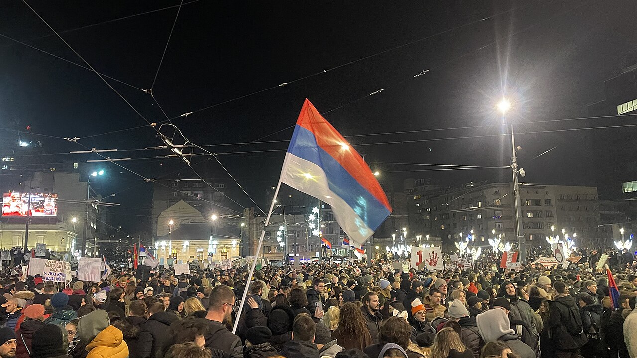 A protest crowd at Slavija Square in Belgrade, with a Serbian flag and a red handprint placard, the symbol of the student movement. The square hosted the Student List final rally on Thursday, when the Archive of Public Gatherings estimated around 150,000 people took part.